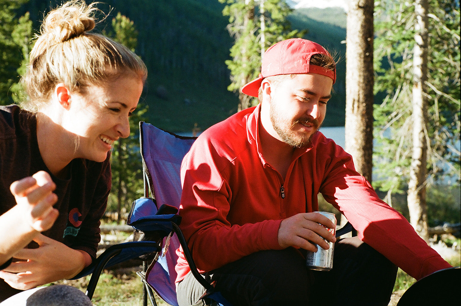 Two campers sit in camping chairs in front of an alpine lake