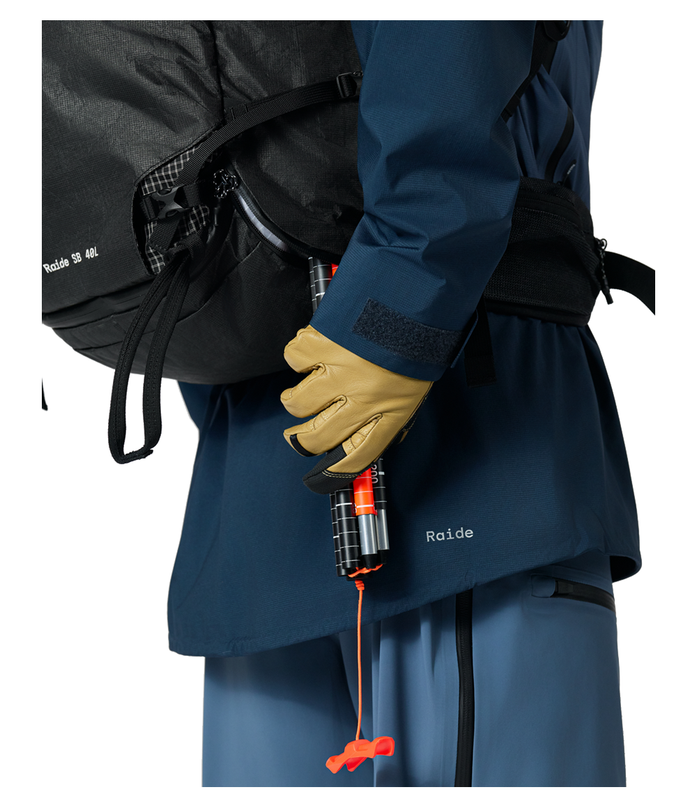 Raide SB 40L Black backcountry ski pack worn on torso showing side-panel avalanche probe access with tan gloves gripping collapsible probe with orange handle and cord