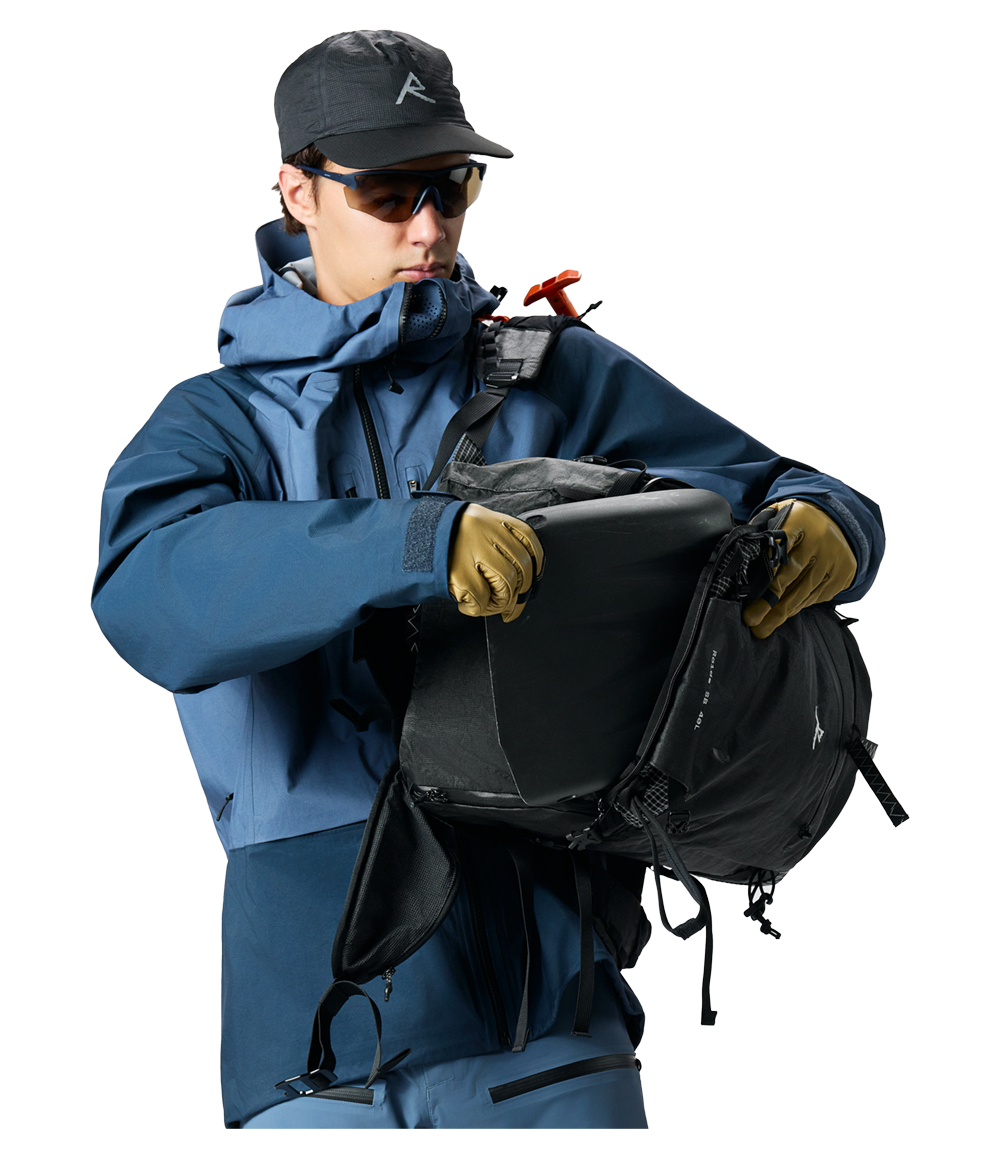 Raide SB 40L backcountry ski pack in Black with back panel access open, showing internal compartment and removable frame, worn by skier in blue jacket and tan gloves with ice tools visible on shoulder straps