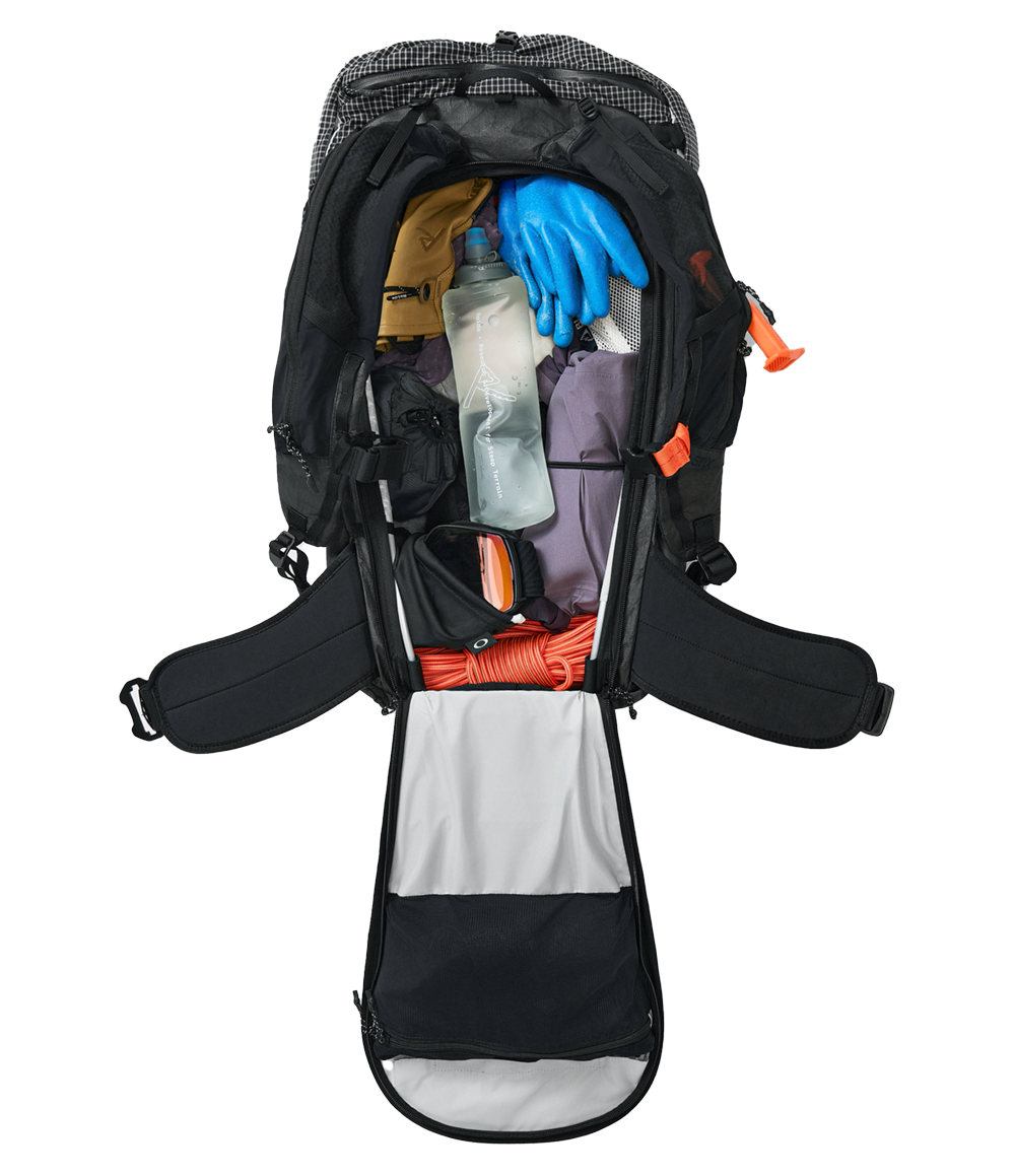 Raide SB 40L Black backcountry ski pack shown with back panel access fully open, revealing main compartment packed with hydration bladder, climbing skins, puffy jacket, and touring essentials, plus padded hip belt and orange safety whistle attachments.