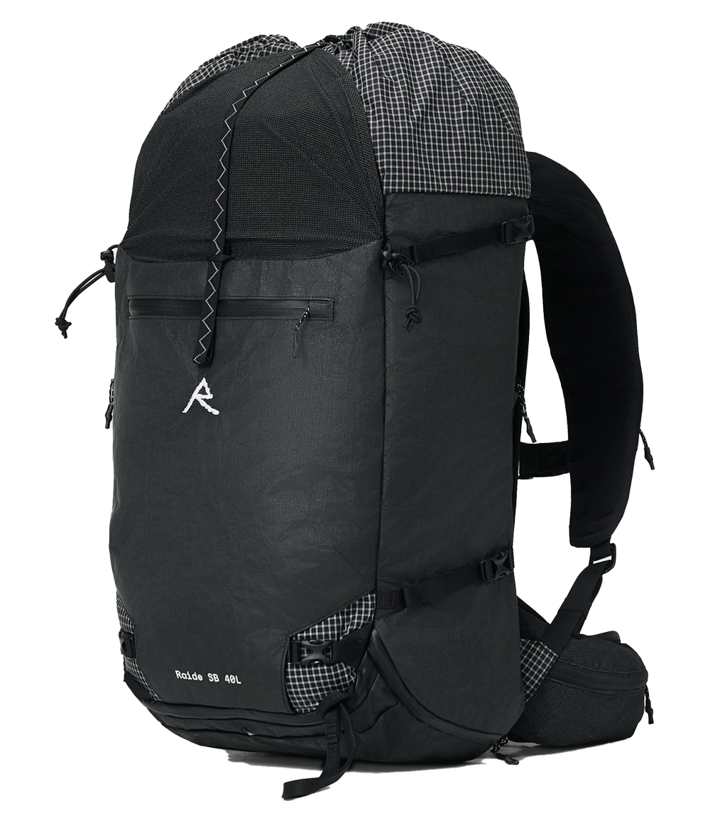 Raide SB 40L backcountry ski pack in Black with extended roll-top compartment, padded shoulder straps, hip belt with zippered pockets, and diagonal ski carry straps, shown loaded with gear including a plaid shirt
