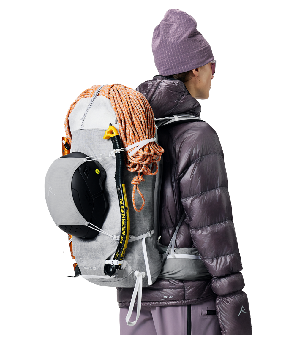 Raide UL 35L backcountry ski pack in light grey technical fabric with orange rope lashing, shown worn with snowboard and avalanche probe attached to exterior carry system